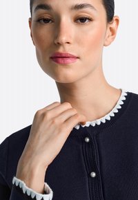 Námornícky pletený kardigán s vlnitým bielym lemom a dvoma ozdobnými perlami na gombíky. Detail výstrihu a ruky pri golieri.