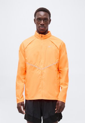 Mężczyzna ubrany w jaskrawo pomarańczową sportową kurtkę zapinaną na zamek i czarne sportowe spodenki, stojący na jednolitym białym tle.