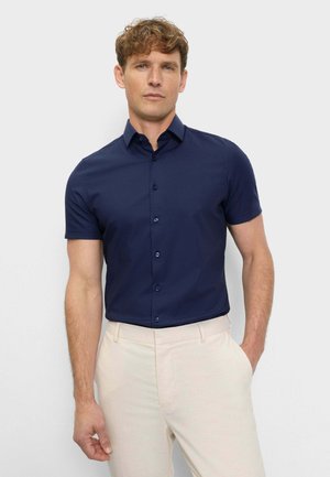 Mann trägt ein eng anliegendes, marineblaues Kurzarm-Hemd mit Knöpfen, in eine hellbeige Hose gesteckt, steht mit einer Hand in der Tasche.