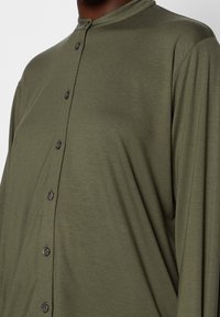 Olijfgroene blouse met lange mouwen, een ronde kraag en zwarte knopen aan de voorkant, gemaakt van een gladde, lichtgewicht stof.