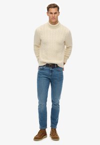 Mand står iført en krämfarvet kabelstrikket sweater, blå jeans, brune læderstøvler og et mørkt bælte mod en lys hvid baggrund.