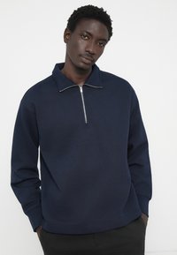 Sudadera con cremallera de color azul marino, fabricada en un material suave, con un alto cuello. Presenta una cremallera plateada y un corte relajado, además de puños y dobladillo acanalados.