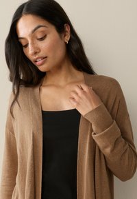 Light brown cardigan with open front, ribbed cuffs, and soft texture, layered over a black top, set against a neutral background.