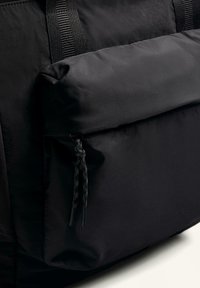 Fekete nylontáska nagy külső zsebbel, fonott zsinórral és lapos pántokkal a hordozáshoz. Simább textúrájú, matt felülettel.