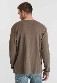 Hombre de espaldas vistiendo una camisa de manga larga marrón y vaqueros azules frente a un fondo claro y liso.