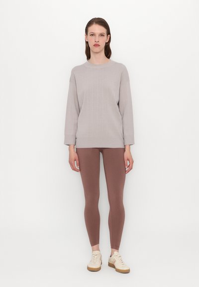 Femeie stând în fața unui perete alb, purtând un pulover din lână, gri deschis, colanți mauve și adidași bej.