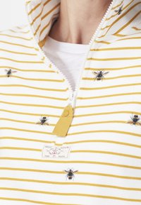Top de cremallera a rayas amarillas y blancas con abejas bordadas, tejido texturizado y una pequeña etiqueta en el escote.