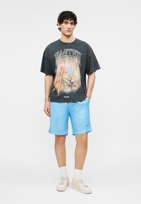Junger Mann steht mit den Händen in den Taschen, trägt ein schwarzes T-Shirt mit Aufdruck, hellblaue Shorts, weiße Socken und beige Turnschuhe.