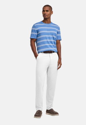 Man standing with hands in pockets wearing blue striped short-sleeve shirt, white pants, brown belt, and brown shoes on plain background.