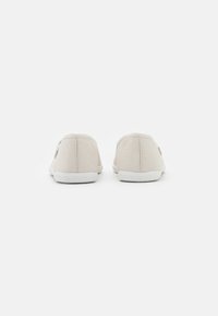 Sapatos slip-on em lona bege com solas de borracha brancas, parte traseira texturizada e detalhes mínimos nas laterais, apresentando um design de bico arredondado.