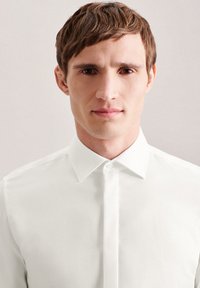 Camicia bianca con colletto a punta e patta frontale; texture liscia e design aderente; il modello ha i capelli corti e uno sfondo neutro.