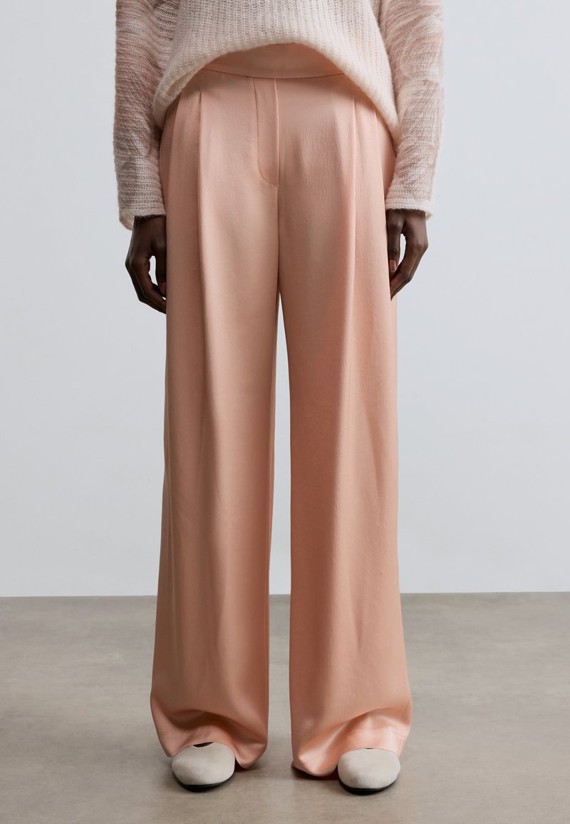 Pantalon large rose en tissu lisse, avec une coupe décontractée et des plis à l'avant. Associé à des chaussures plates de couleur claire.