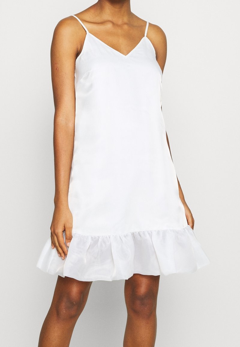 Femme portant une robe blanche en satin sans manches avec des bretelles fines et un ourlet volanté, debout devant un fond clair uni.