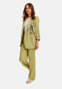 Světle zelený oversize blazer s odpovídajícími kalhotami, doplněný květinovou šálou uvázanou na rukávu a béžovými teniskami. Hladká textura.