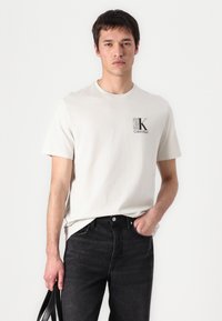 Bílá krátká tričko s krátkým rukávem a černým logem "CK" a textem "Calvin Klein". Nosené s tmavými džíny, ukazující uvolněný střih. Hladká bavlněná textura.