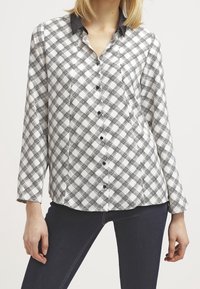 Kobieta ubrana w koszulę w czarno-białą kratę z długim rękawem i czarnym kołnierzykiem, w komplecie z ciemnoniebieskimi dopasowanymi spodniami.