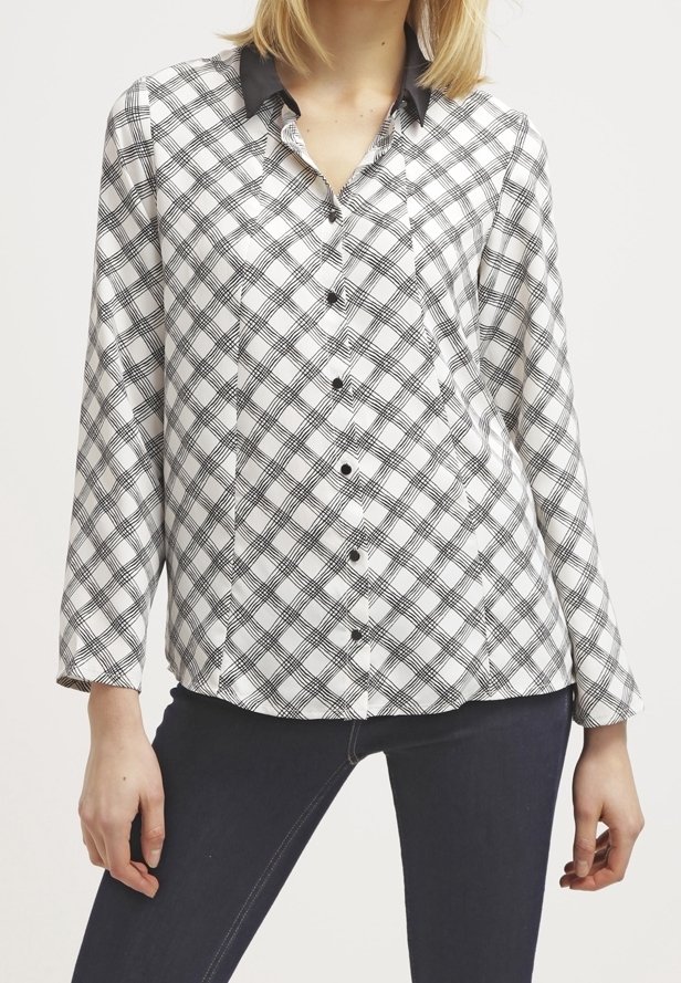 Kobieta ubrana w koszulę w czarno-białą kratę z długim rękawem i czarnym kołnierzykiem, w komplecie z ciemnoniebieskimi dopasowanymi spodniami.