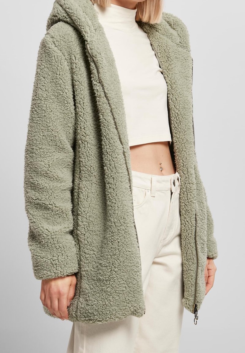 Mujer vistiendo una chaqueta de felpa verde claro con cremallera, un jersey corto blanco de cuello alto, y pantalones blancos de tiro alto, mostrando un piercing en el ombligo.