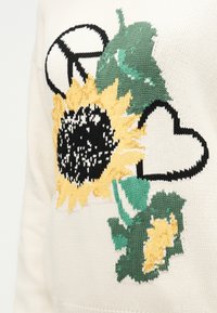 Πλεκτός ηλιοτρόπιο με πράσινα φύλλα, μαύρο σύμβολο ειρήνης και περίγραμμα μαύρης καρδιάς σε κρεμ ύφασμα.