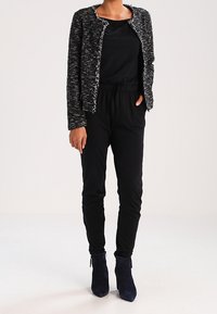 Blazer noir texturé avec des bords effilochés, associé à un haut noir et un pantalon fuselé. Le modèle porte des bottines bleu marine. Look simple et taillé sur mesure.