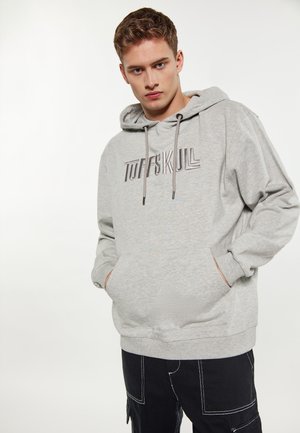 Jonge man die een lichtgrijze hoodie draagt met de tekst "TUFFSKULL" en een zwarte broek met witte steken, handen in de zakken van de hoodie, neutrale achtergrond.