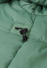 Tessuto di giacca verde gonfia con una texture liscia; presenta un cordino regolabile e un'etichetta con il marchio sul petto.