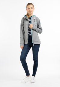 Grå softshell-jacka med en ljusblå dragkedja och huva, med snoddar. Kombinerad med mörka jeans och vita sneakers. Randig skjorta under.