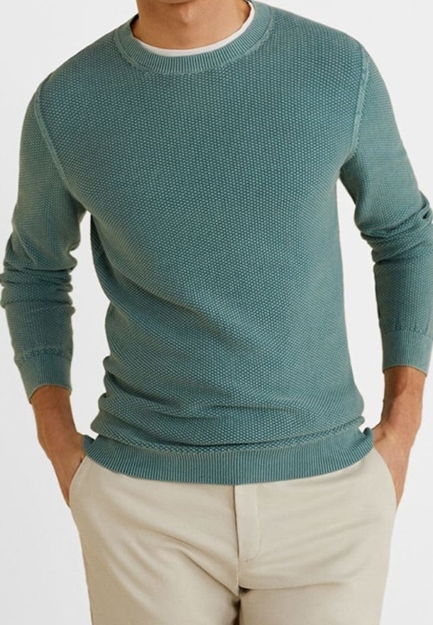 Homme portant un pull en tricot texturé bleu canard et un pantalon beige, les mains dans les poches, recadré sous le menton.