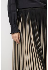 Woman wearing black long-sleeve blouse with smocked cuffs and a high-waisted pleated skirt with black and beige vertical stripes.