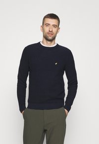 Pulover tricotat în dungi bleumarin, cu guler rotund, mâneci lungi și un mic logo galben pe piept, asortat cu pantaloni de culoare verde măslin.