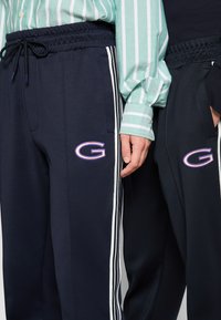 Joggers navy avec des rayures latérales blanches et un logo "G" brodé, taille élastique et poches. Tissu lisse avec une coupe décontractée.