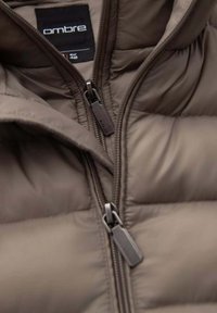 Bruine pufferjas met een hoge kraag, opvallende stiksels, dubbele ritsen en een label met het merk "Ombre" en maat 48.