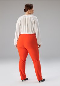 Weiße leichte Bluse mit langen Ärmeln, kombiniert mit leuchtend roten, figurbetonten Hosen und schwarzen spitzen Absatzschuhen, von hinten gesehen.