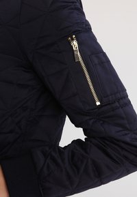 Chaqueta acolchada de color marino con un bolsillo con cremallera en la manga izquierda, que presenta una cremallera en tono dorado y puños acanalados.