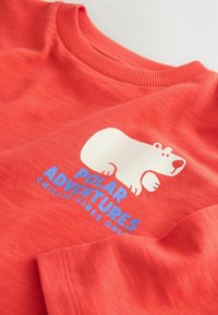 Maglietta rossa con un orso polare in stile cartone animato sopra il testo blu "Avventure Polari Solo Vibrazioni Rinfrescanti" nell'area del petto.