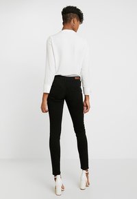 Svarta smala jeans med medelhög midja och två bakfickor. Vit långärmad topp med en texturerad yta. Modell som bär klacksandaler.