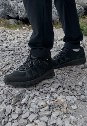 Personne portant des chaussures de randonnée noires et un pantalon noir, debout sur un sol rocheux en extérieur.
