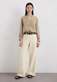 Beige Leinenbluse mit Kragen und Knopfleiste, kombiniert mit crèmefarbenen weit geschnittenen Hosen. Schwarzer Gürtel mit goldener Schnalle und flachen schwarzen Sandalen.