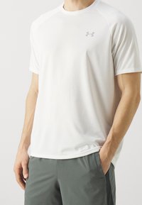 Camiseta deportiva blanca que absorbe la humedad, de manga corta y cuello redondo. Presenta un logo gris sutil en el pecho.