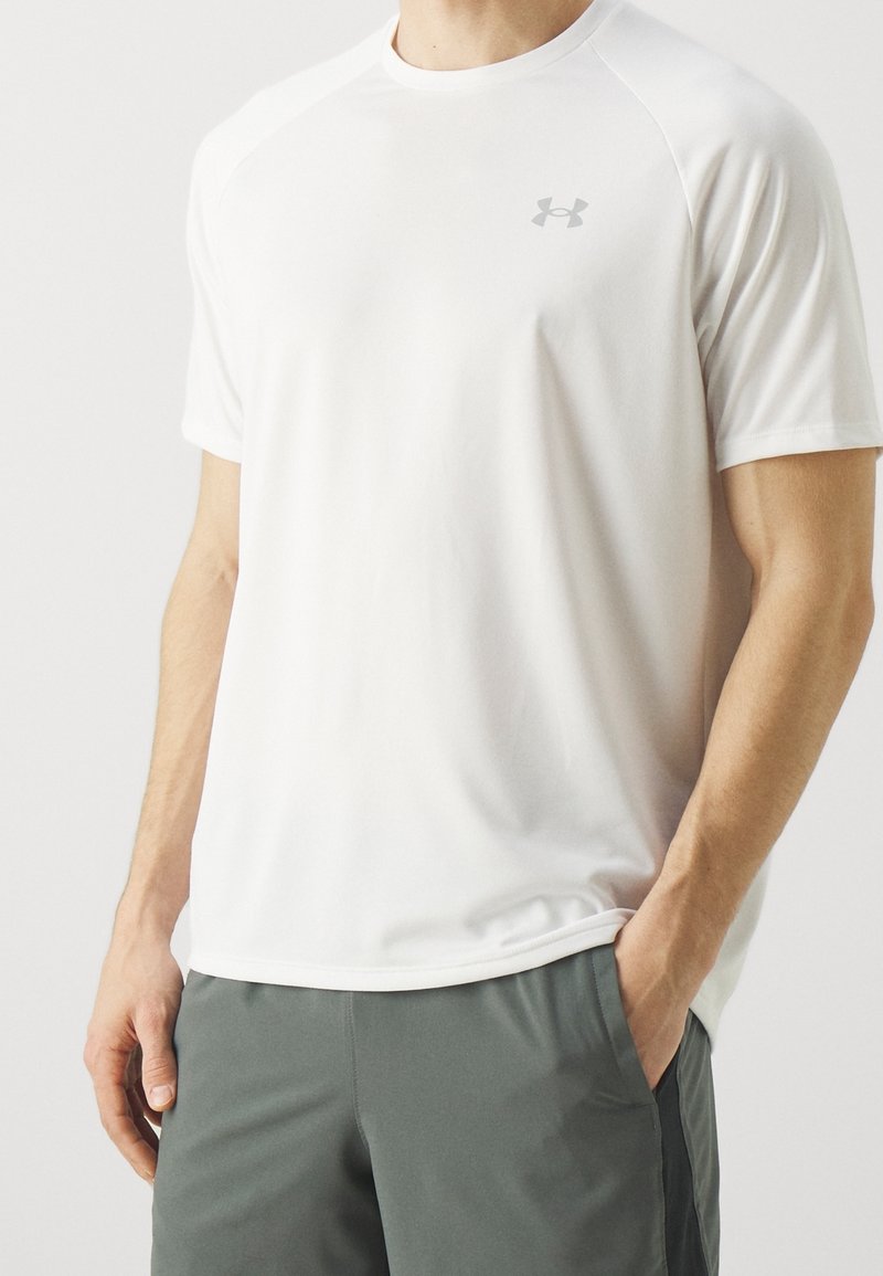 Camiseta deportiva blanca que absorbe la humedad, de manga corta y cuello redondo. Presenta un logo gris sutil en el pecho.