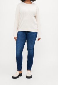 Maglione beige lavorato a maglia con motivo testurizzato, abbinato a jeans slim-fit blu scuro e scarpe bianche con suole nere e dettagli in frangia.