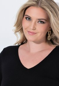 Schwarzer, strukturierter V-Ausschnitt-Pullover kombiniert mit goldenen geometrischen Ohrringen. Weicher, mittelschwerer Stoff. Das Model hat welliges, blondes Haar und ein neutrales Make-up.