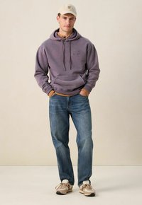 Jonge man met een beige pet, paarse hoodie over een bruine shirt, blauwe jeans en beige sneakers, staand met zijn handen in zijn zakken.