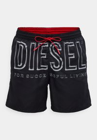 Mustad ujumispüksid punase elastse vöökohaga, millel on suur hõbedane "DIESEL" logo ja tekst "FOR SUCCESSFUL LIVING" vastanduvates õmblustes.