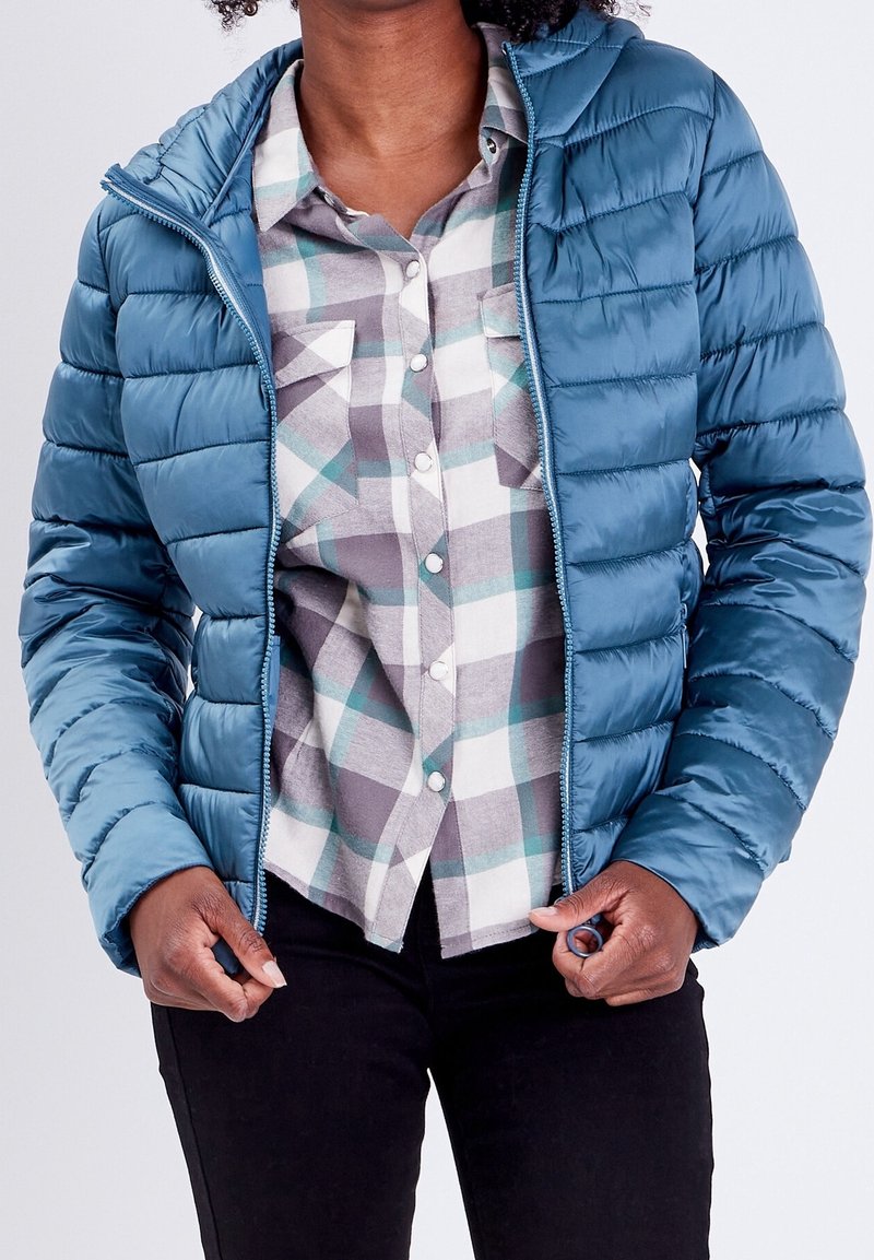 Personne portant une doudoune bleue ouverte sur une chemise à carreaux grise et blanche boutonnée, tenant les bords de la veste, vêtue d’un pantalon noir.