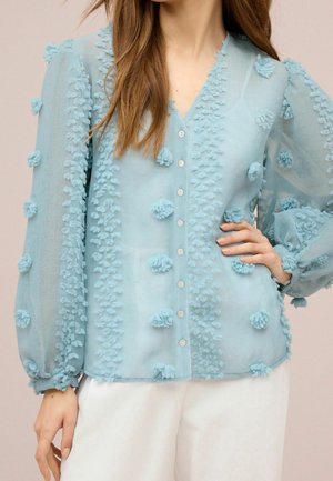 Femme portant un chemisier bleu clair transparent avec des détails pompons texturés et un pantalon blanc, main sur la hanche, devant un fond uni.