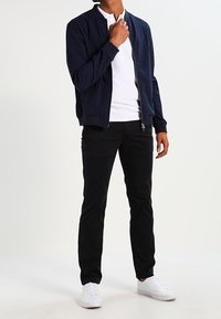 Námořnicky modrá bomber bunda přes bílou polokošili, doplněná černými úzkými kalhotami a bílými teniskami, vytvářející ležérní outfit.