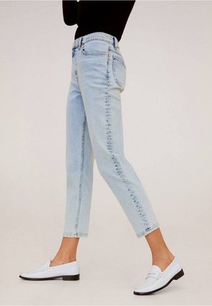 Personne portant un jean court bleu clair, un haut noir et des mocassins blancs, debout de profil sur un fond uni.