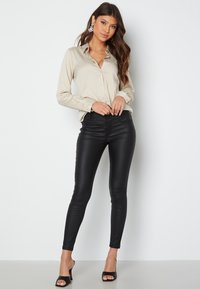 Beige overhemd met knopen en een kraag, gecombineerd met glanzende zwarte skinny broeken. Het model draagt zwarte hoge hakken en decoratieve oorbellen.