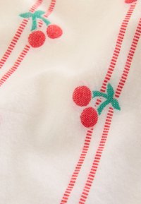 Bílá látka s motivem červených třešní a zelených stonků, doplněná svislými červenými tečkovanými čarami.